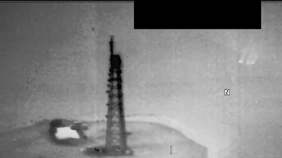 Infrarrojo en grises: una torre de celosía oscura sobre un islote, con un punto blanco brillante a su izquierda y, arriba a la derecha, un grupo de puntos claros con estela.