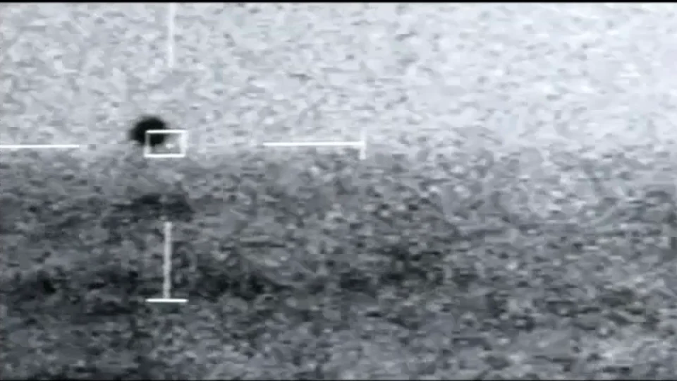 Infrarrojo en grises grabado de una pantalla: cielo claro arriba, mar oscuro abajo y, justo sobre la línea entre ambos, una mancha negra redonda con un recuadro blanco y la retícula.
